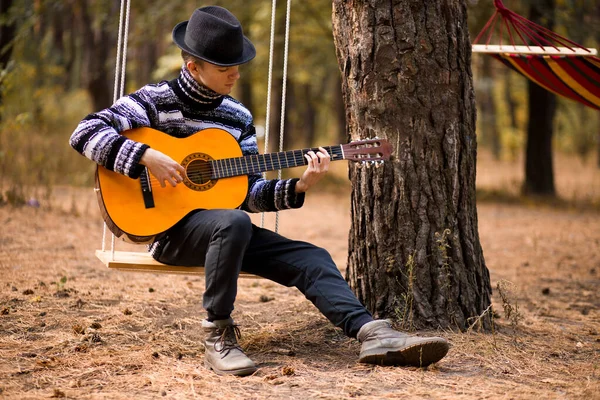 Joven hombre atractivo guapo en suéter tocando la guitarra en el bosque sentado en swing. Hombre casual, estilo de vida . — Foto de Stock