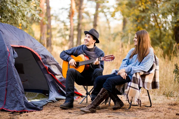 Retrato de pareja joven sentada en silla de campo con guitarra cerca de la tienda de campaña. Tiempo juntos en armonía con la naturaleza lejos del bullicio de la ciudad — Foto de Stock