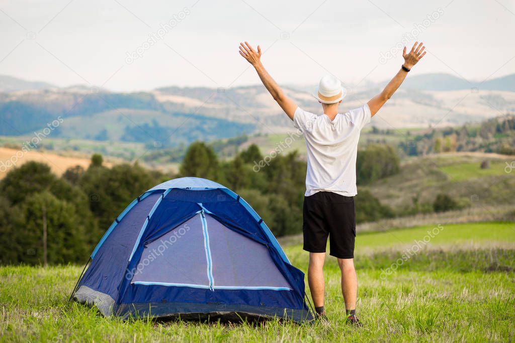 Young man with hands up camping tent facing amazing views of mountain