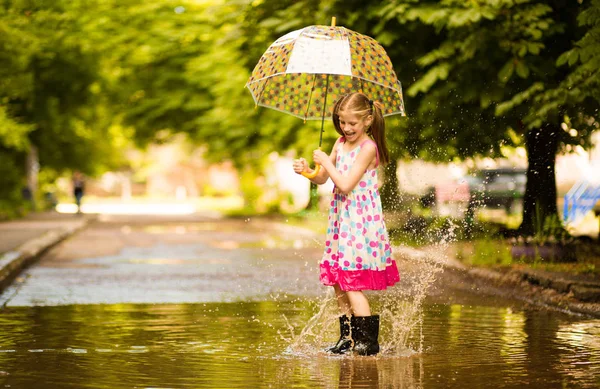 Feliz niña divertida con paraguas saltando sobre charcos en botas de goma y riendo —  Fotos de Stock