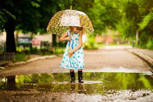 Menina garoto engraçado feliz com guarda-chuva pulando em poças em botas de borracha e rindo — Fotografia de Stock