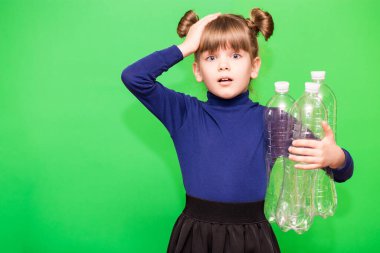 Şok olmuş yüzü ve eli plastik şişelerle dolu ve yeşil arka planda izole edilmiş kameraya şaşkın şaşkın bakan küçük kız.