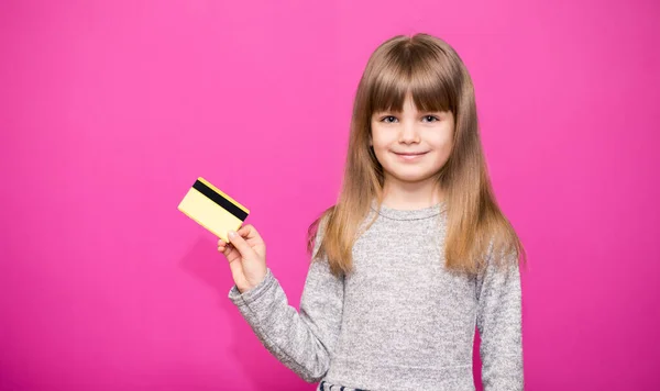 Mutlu küçük kız elinde yeni bir banka kartı tutuyor ve pembe arka planda alışverişe gitmeye hazır.