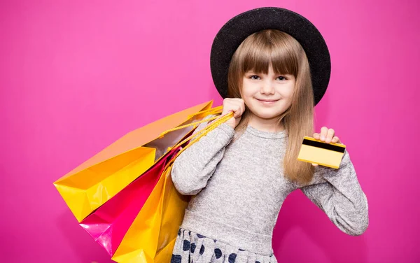 戴着黑色帽子的漂亮小女孩拿着购物袋和信用卡 背景是粉色的 — 图库照片