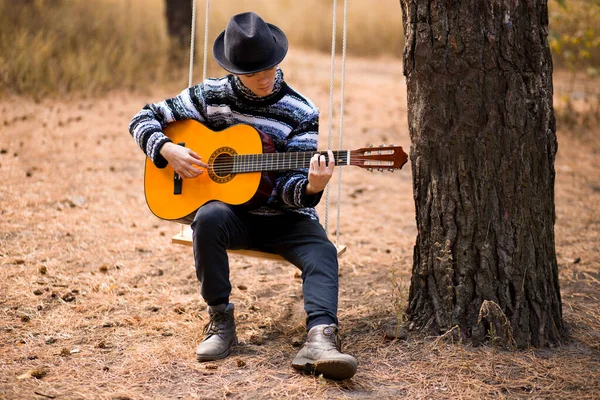 young handsome attractive man in sweater and black hat playing guitar in forest sitting on swing. Casual man, life style.