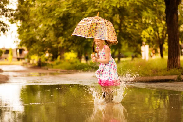 ゴムブーツと水たまりの水たまりに飛び乗る傘を持つ幸せな面白い子供の女の子と水玉のドレスと笑い — ストック写真