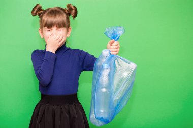 Küçük çevre bilimci kız çöp torbasını plastikle tutuyor ve burnunu yeşil arka planda izole ediyor. Gezegenin plastik kirliliği