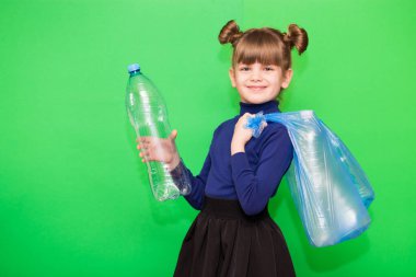 Plastik şişelerle dolu çöp torbası tutan küçük pozitif bir çevreci kız ve yeşil arka planda izole edilmiş bir parmak gösteriyor. Ekolojik çevre problemi tüketimi. Plastik tüketimini azaltmak kârlıdır.