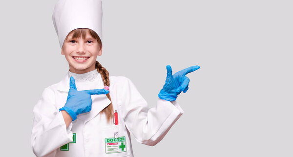 Smiling little girl in medical uniform and blue gloves point to right on free space looking at camera isolated on grey background. Sterility and disinfection concept