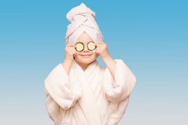 Beyaz önlüklü, maskeli, deri yüzlü ve mavi arka planda gözlerinde salatalık olan komik küçük kız. Spa cilt bakımı