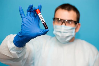 Mavi arka planda izole edilmiş koruyucu maskeli doktor ve eldivenlerde koronavirüs için test edilmiş kan örneği var. Coronavirus COVID 19 için hastaların kanını test ediyoruz..
