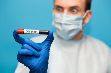 Mavi arka planda izole edilmiş koruyucu maskeli doktor ve eldivenlerde koronavirüs için test edilmiş kan örneği var. Coronavirus COVID 19 için hastaların kanını test ediyoruz..
