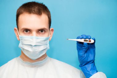 Koruyucu maskeli ve eldivenli yakışıklı doktor mavi arka planda 40 derece izole tıbbi termometre tutuyor. Sıcaklık, grip veya yetersiz teşhis ekipmanları