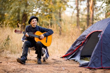 Siyah şapkalı gülümseyen genç adam sonbahar ormanlarında turistik çadırın yanında oturup gitar çalıyor.