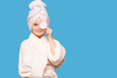 Başında havluyla gülümseyen güzel bir kız çocuğu pamuk bloknotla deriyi temizliyor mavi arka planda izole edilmiş temizleyici ile. Mutlu kız makyajını sil ve kameraya bak. Sağlıklı, temiz cilt bakımının keyfini çıkar.