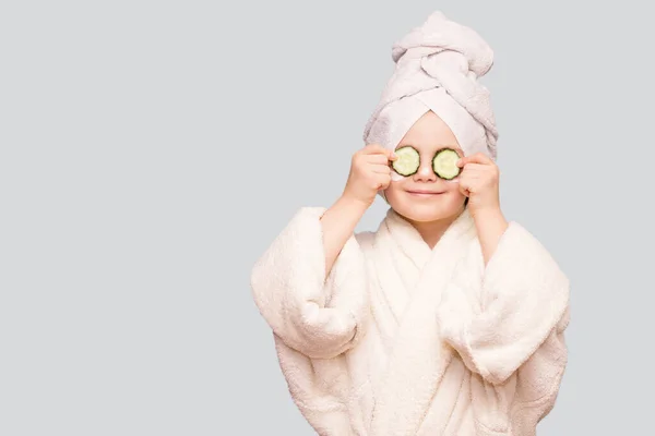 Beyaz önlüklü ve maske takan komik küçük bir kız ve beyaz arka planda gözlerinde salatalık var. Spa cilt bakımı
