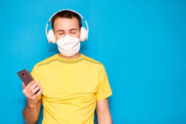 Tıbbi maskeli ve kulaklıklı yakışıklı sükunetli adam mavi arka planda izole edilmiş müzik dinliyor. Dışarıdan gelen bilgileri filtrele