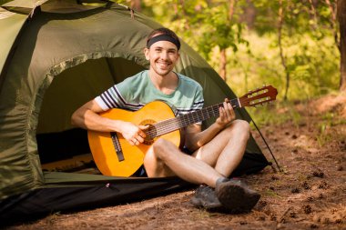 Mutlu genç adam kamp yapıyor ve ormanın günbatımına karşı dinlenmek için gitar enstrümantal bir müzik çalıyor. Yaban hayatı hayat tarzı konsepti.