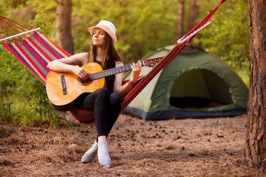 Şapkalı kadın gitar çalıyor ve ormanın ortasında hamakta dinleniyor. Yavaş yaşam konsepti. Hippi..