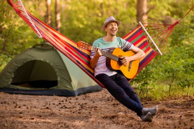 Şapkalı genç beyaz adam gitar çalıyor ve hafta sonu kampta hamakta sallanıyor.