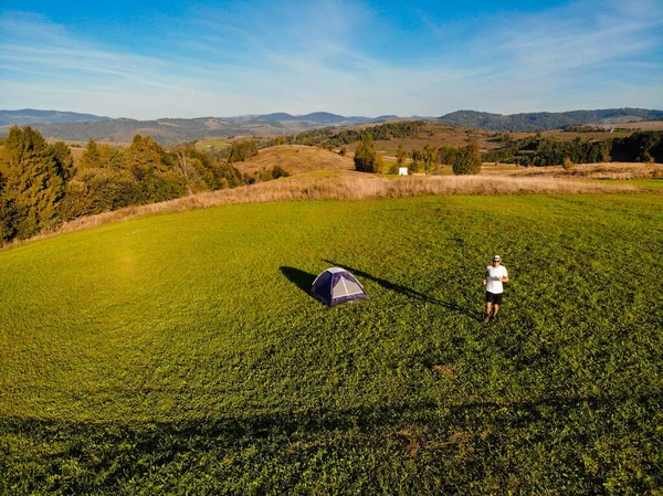 Aerial view of young man run by meadow near tent with mountains background. Exercise wellbeing concept