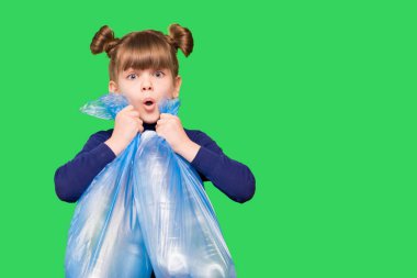 Kafası karışmış, komik suratlı, yeşil arka planda plastik şişelerle dolu çöp torbaları tutan küçük bir çevreci kız. Ekoloji çevre sorunu tüketimi