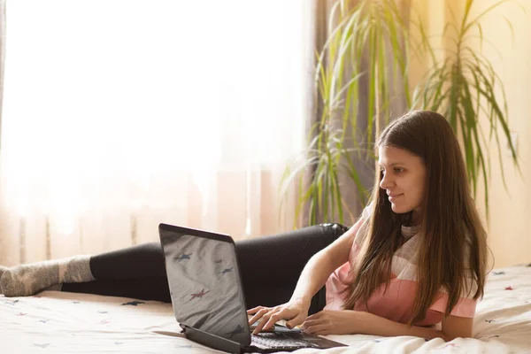 Young woman working on laptop surfing net at home and smiling while sitting on bed at home. Distance work. Telework. Stay home