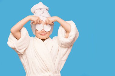 Başında havluyla gülümseyen güzel bir kız çocuğu pamuk bloknotla deriyi temizliyor mavi arka planda izole edilmiş temizleyici ile. Mutlu kız makyajını sil ve kameraya bak. Sağlıklı, temiz cilt bakımının keyfini çıkar.