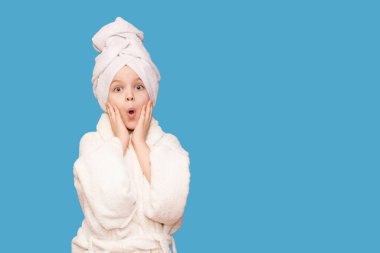 Güzellik tedavisinden sonra saç havlusu ve beyaz önlük giyen kaplıca güzeli kız. Şok olmuş yüzü ve mavi arka planda izole edilmiş mükemmel cildi olan güzel bir kız çocuğu.