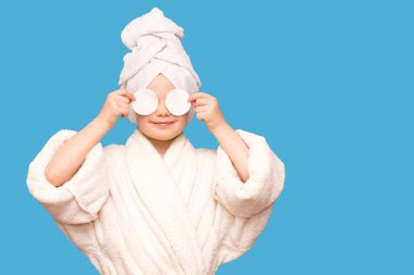 Başında havluyla gülümseyen güzel bir kız çocuğu pamuk bloknotla deriyi temizliyor mavi arka planda izole edilmiş temizleyici ile. Mutlu kız makyajını sil ve kameraya bak. Sağlıklı, temiz cilt bakımının keyfini çıkar.