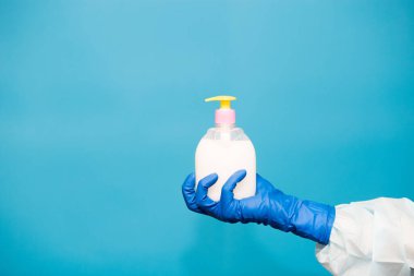 Mavi eldiven giyen kişi mavi arka planda izole edilmiş sabun şişesini tutuyor. Eller hijyen virüsü salgını önlüyor.