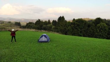 Genç mutlu kadının hava aracı görüntüsü çadırın yanında duruyor ve ellerini kaldırıyor manzaralı dağ günbatımının arka planında.