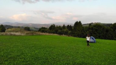 Genç mutlu kadının hava aracı görüntüsü çadırın yanında duruyor ve ellerini kaldırıyor manzaralı dağ günbatımının arka planında.