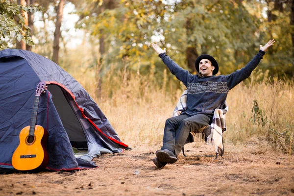 Smiling young man in black hat sitting near touristic tent and playing guitar in autumn forest