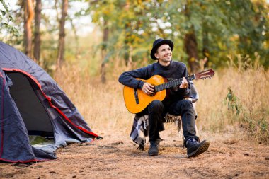 Siyah şapkalı gülümseyen genç adam sonbahar ormanlarında turistik çadırın yanında oturup gitar çalıyor.