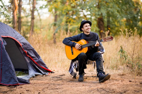 Smiling young man in black hat sitting near touristic tent and playing guitar in autumn forest