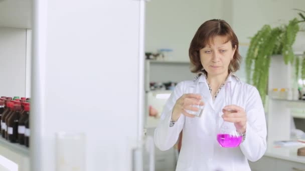 Químico no laboratório. Closeup de mulher química olhando para líquido em frasco de vidro no laboratório. Mulher química analisa fluido químico em frasco de laboratório rosa — Vídeo de Stock