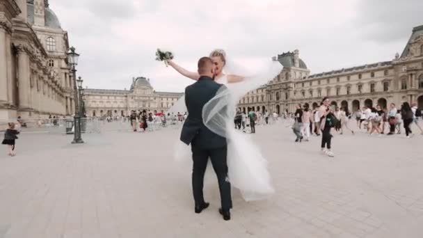 프랑스 파리에 있는 루브르 박물관 근처에서 행복 한 부부가 있습니다. 신랑은 신부를 팔로 비틀어 댄다. 베일을 흔들고 있는 신부 — 비디오