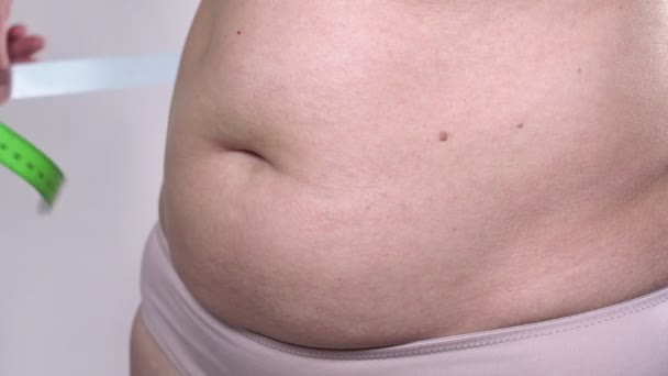 Žena středního věku s povislou pletí definuje své velké břicho měřicí páskou na bílém pozadí. Vedlejší plán. — Stock video
