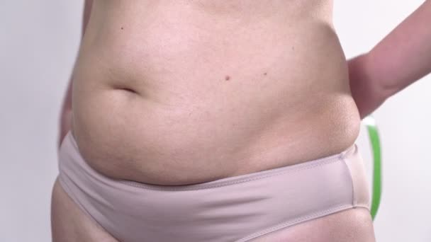 Een vrouw van middelbare leeftijd met een slappe huid definieert haar grote buik met een meetlint, op een witte achtergrond. Zijplan. — Stockvideo