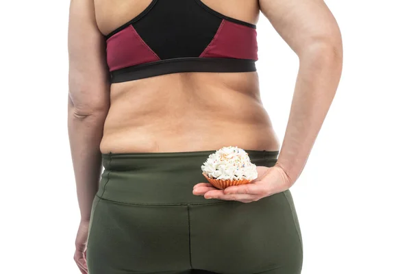 Een vrouw van middelbare leeftijd, zwaarlijvige en slappe huid, met een cupcake in haar hand verborgen achter haar rug, op een witte achtergrond, close-up. — Stockfoto