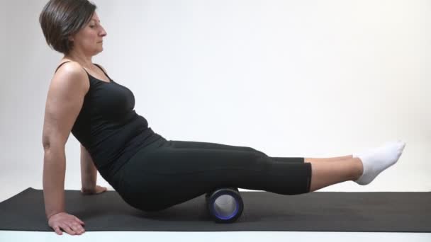 Een vrouw van middelbare leeftijd op een gymnastiekmat met myofasciale roller doet een oefening op haar heupen op een witte achtergrond. — Stockvideo