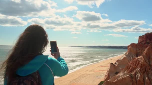 Молода дівчина турист на пляжі біля океану з телефоном в руці зробить фотографії ландшафту навколо нього . — стокове відео