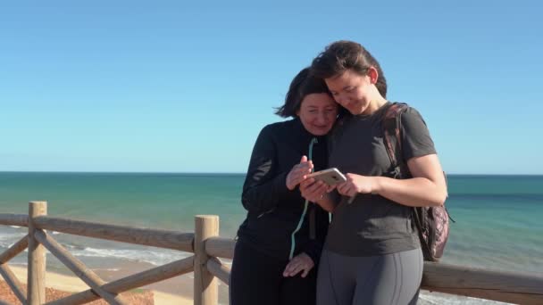 Mulher de meia-idade bem sucedida e feliz com uma jovem, em uma rocha perto do oceano ri do que ele viu no telefone. Portugal. Vilamoura. Mãe com filha . — Vídeo de Stock