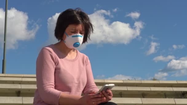 Женщина средних лет в защитной медицинской маске от вируса и аллергии сидит на скамейке с телефоном и смартфоном в руках. . — стоковое видео