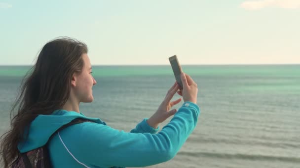 Jovem turista na praia junto ao oceano com um telefone na mão vai tirar fotos da paisagem ao seu redor . — Vídeo de Stock