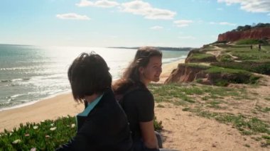 Başarılı, orta yaşlı bir kadın, genç bir kızla birlikte okyanusun kenarındaki bir uçurumda yorgun bir şekilde bir taşın üzerine oturmuş. Portekiz. Vilamoura. Anne ve kızı..