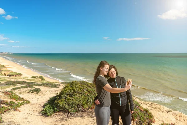 Dwie sportowe dziewczyny, na skalistym brzegu, zrobić zdjęcie selfie, odnoszący sukcesy biznesmeni sortuje Ocean Atlantycki Portugalia, Falesia Algarve. — Zdjęcie stockowe