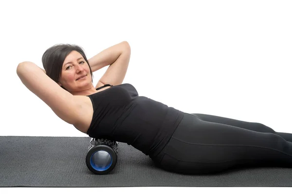 En medelålders kvinna med slapp hud på en gymnastikmatta med myofascial roller gör en övning på ryggen på en vit bakgrund. — Stockfoto