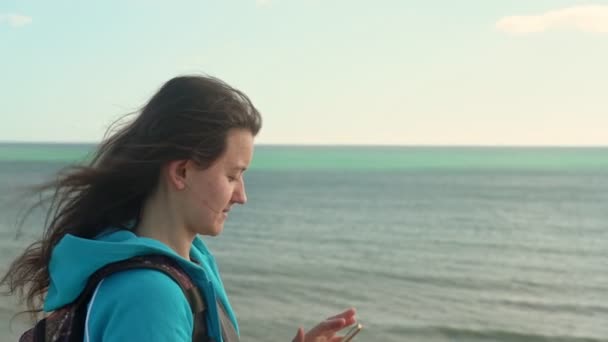 Jovem turista na praia junto ao oceano com um telefone na mão vai tirar fotos da paisagem ao seu redor . — Vídeo de Stock